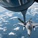 An aerial refueling operation is captured from the perspective of a tanker aircraft, showing its refueling boom extended toward an F-16 Fighting Falcon fighter jet. The F-16 is marked with 'PA' and 'ANG' in yellow lettering, indicating it belongs to the Pennsylvania Air National Guard. The aircraft are flying at altitude above a partially cloud-covered landscape.