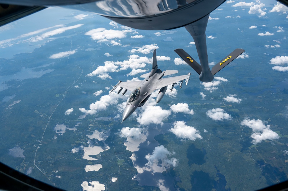 An F-16 Fighting Falcon fighter jet is approaching a KC-135 Stratotanker for aerial refueling at high altitude. The refueling boom of the tanker aircraft is visible in the foreground, marked with 'PA ANG' indicating Pennsylvania Air National Guard. The aircraft are flying over a landscape with scattered cumulus clouds, lakes, and green terrain visible far below.
