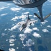 An F-16 Fighting Falcon fighter jet is approaching a KC-135 Stratotanker for aerial refueling at high altitude. The refueling boom of the tanker aircraft is visible in the foreground, marked with 'PA ANG' indicating Pennsylvania Air National Guard. The aircraft are flying over a landscape with scattered cumulus clouds, lakes, and green terrain visible far below.