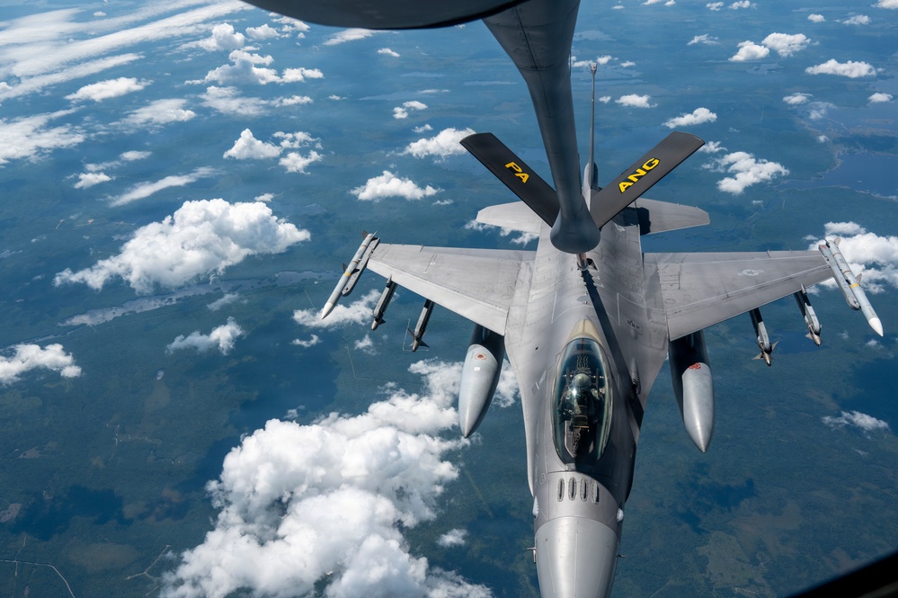 An F-16 Fighting Falcon fighter jet is conducting an aerial refueling operation with a tanker aircraft, visible at the top of the frame. The fighter jet bears markings 'PA' and 'ANG' on its tail fins, indicating it belongs to the Pennsylvania Air National Guard. The aircraft is flying at altitude above scattered cloud cover over what appears to be a rural landscape.