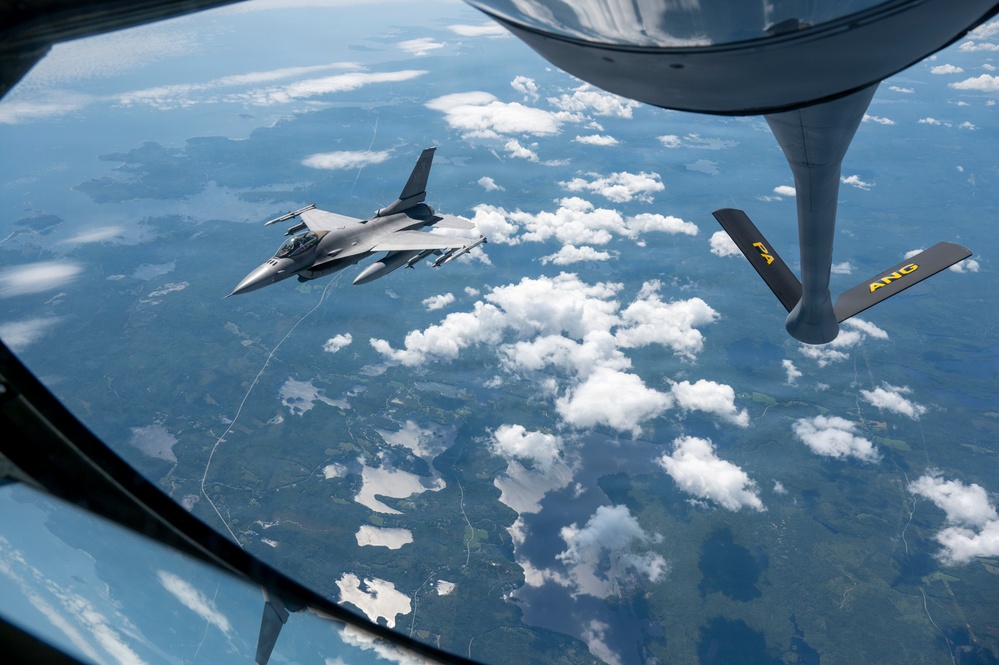 An aerial refueling operation is captured from the boom operator's window of a tanker aircraft, showing an F-16 Fighting Falcon connected via a refueling boom. The tanker's boom and stabilizer fins are visible in the foreground, with markings 'PA' and 'ANG' indicating Pennsylvania Air National Guard affiliation. The aircraft are flying over a landscape of forests, lakes, and scattered cumulus clouds.