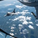 An aerial refueling operation is captured from the boom operator's window of a tanker aircraft, showing an F-16 Fighting Falcon connected via a refueling boom. The tanker's boom and stabilizer fins are visible in the foreground, with markings 'PA' and 'ANG' indicating Pennsylvania Air National Guard affiliation. The aircraft are flying over a landscape of forests, lakes, and scattered cumulus clouds.