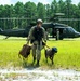 A military service member in camouflage uniform walks away from a United States Army UH-60 Black Hawk helicopter that has landed in a grassy clearing surrounded by pine trees. The operator is accompanied by a military working dog (MWD) equipped with tactical gear including protective goggles (doggles) and a vest, while carrying a tan duffel bag. Additional personnel can be seen near the helicopter in the background, suggesting a training exercise or operational insertion.