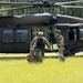A U.S. Army UH-60 Black Hawk helicopter is landed on a grassy field surrounded by pine trees. Two soldiers in combat gear are disembarking from the aircraft, carrying equipment bags, while a pilot remains visible in the cockpit. The scene appears to depict a training or operational insertion exercise.