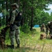 Military personnel in camouflage uniforms are conducting what appears to be a field training exercise in a wooded area. One soldier in the foreground is handling a military working dog (MWD) while other personnel in the background are managing tactical gear and equipment. The setting includes pine trees, open grassland, and portable toilets visible in the background, suggesting a training base or exercise area.