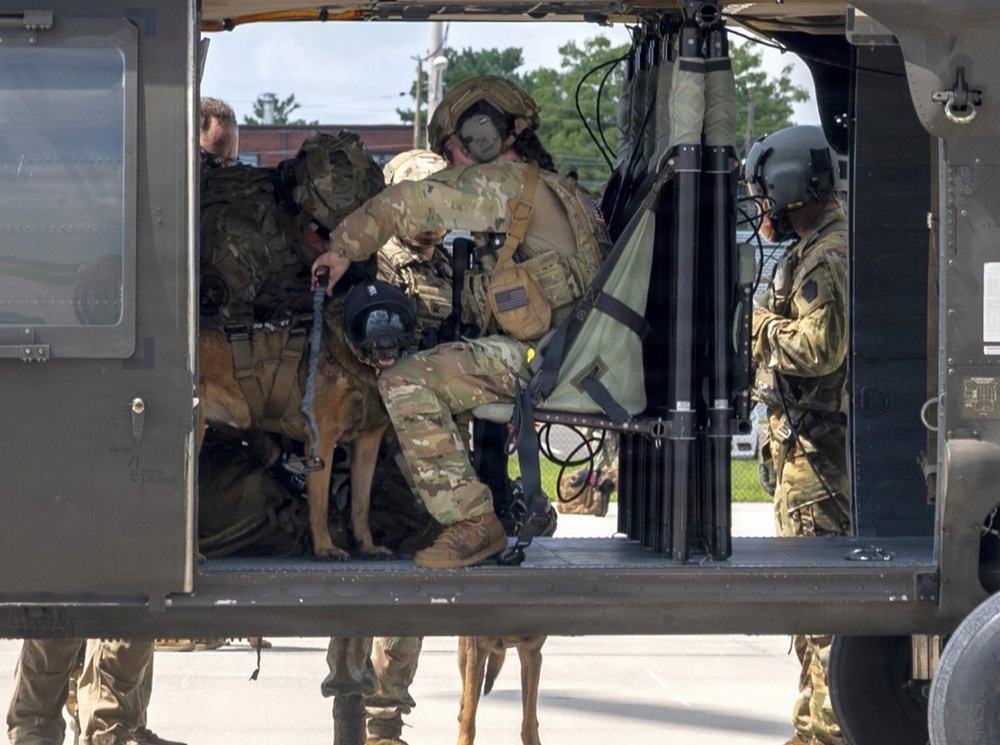 Military personnel in full combat gear are loading or unloading from what appears to be a UH-60 Black Hawk helicopter on a tarmac. A military working dog (MWD), visibly wearing a canine tactical vest or harness, is being handled by one of the soldiers inside the aircraft. The scene depicts a special operations or K9 unit conducting helicopter insertion/extraction training or an operational deployment.