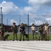 A group of approximately nine military or law enforcement personnel in tactical/camouflage gear are lined up in front of a chain-link fence topped with barbed wire. Several members of the group are accompanied by military working dogs (K9 units). The setting appears to be a secured facility or military installation, photographed on a partly cloudy day.