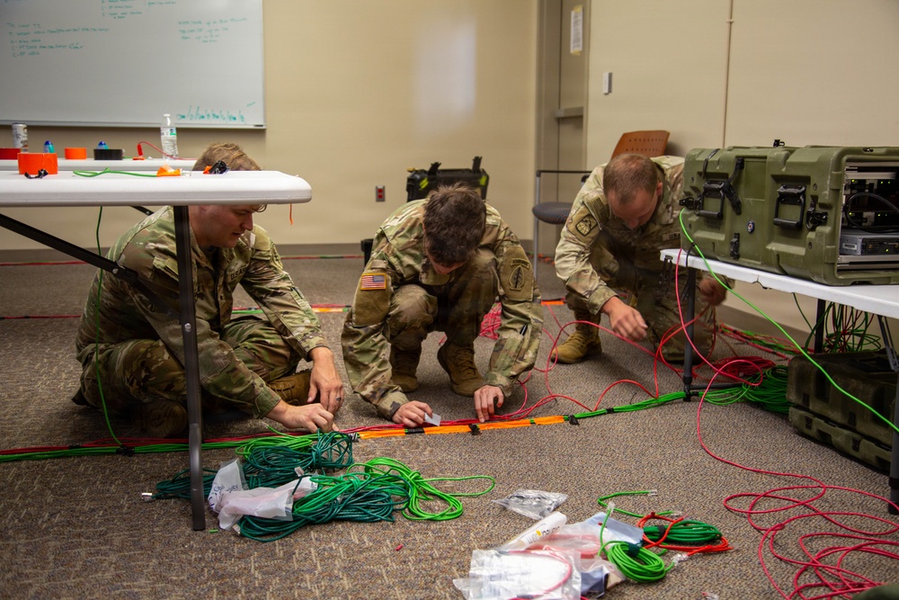Three U.S. Army soldiers in MultiCam/OCP camouflage uniforms are working on the floor of what appears to be a classroom or training room, organizing and routing cables. They are handling multiple colored cables (green, red, and orange) alongside military-grade equipment cases. The setting suggests a communications or signal systems setup exercise or training event.
