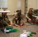 Three U.S. Army soldiers in MultiCam/OCP camouflage uniforms are working on the floor of what appears to be a classroom or training room, organizing and routing cables. They are handling multiple colored cables (green, red, and orange) alongside military-grade equipment cases. The setting suggests a communications or signal systems setup exercise or training event.