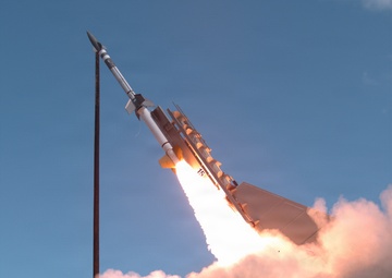 The image captures a missile or rocket at the moment of launch, rising at an angle against a clear blue sky. A large plume of white and pink-tinted exhaust smoke billows from the base of the launch platform. The missile appears to be a surface-to-air or tactical missile system mounted on a launch rail or vehicle.