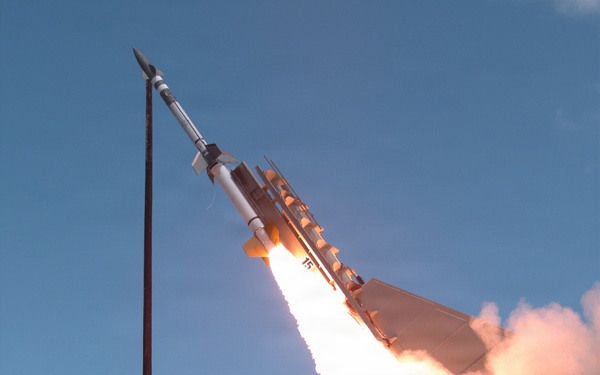 The image captures a missile or rocket at the moment of launch, rising at an angle against a clear blue sky. A large plume of white and pink-tinted exhaust smoke billows from the base of the launch platform. The missile appears to be a surface-to-air or tactical missile system mounted on a launch rail or vehicle.