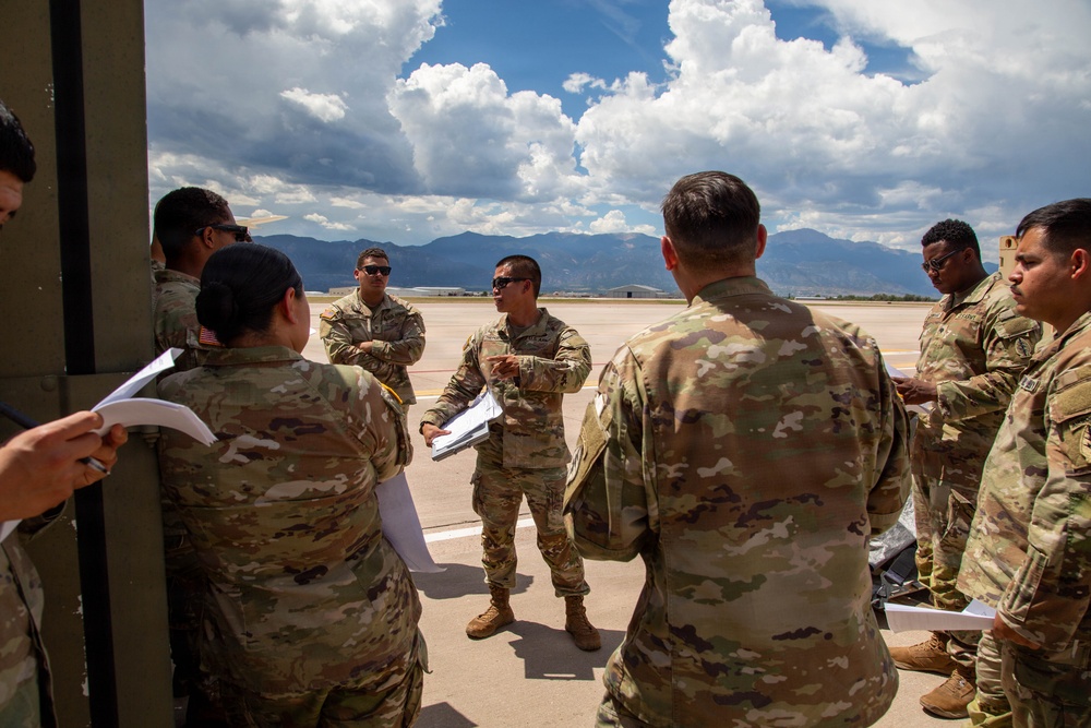 A group of military personnel in OCP (Operational Camouflage Pattern) uniforms are gathered on what appears to be a flight line or tarmac at a military airfield. One soldier in the center appears to be conducting a briefing while holding a clipboard, with mountains and cumulus clouds visible in the background. The setting suggests a pre-mission or operational briefing at a base likely located in a mountainous region.
