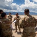 A group of military personnel in OCP (Operational Camouflage Pattern) uniforms are gathered on what appears to be a flight line or tarmac at a military airfield. One soldier in the center appears to be conducting a briefing while holding a clipboard, with mountains and cumulus clouds visible in the background. The setting suggests a pre-mission or operational briefing at a base likely located in a mountainous region.