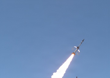 A multi-stage sounding rocket is captured mid-launch, ascending at an angle against a clear blue sky with a long, bright exhaust plume trailing behind it. The launch appears to originate from a small ground facility with support structures visible below. Large billowing white smoke clouds are expanding at the base of the launch site.