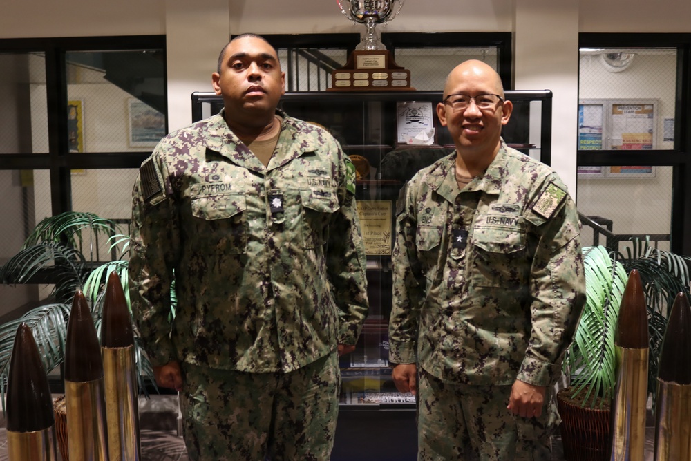 Two U.S. Navy personnel in Navy Working Uniform (NWU Type III) camouflage are posing together indoors. The individual on the left has a name tape reading 'PYFROM' and the one on the right appears to have a name tape reading 'ENG', both displaying 'U.S. NAVY' chest tapes. They are standing in what appears to be a military office or lobby area with trophies and display cases visible in the background.