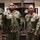 Two U.S. Navy personnel in Navy Working Uniform (NWU Type III) camouflage are posing together indoors. The individual on the left has a name tape reading 'PYFROM' and the one on the right appears to have a name tape reading 'ENG', both displaying 'U.S. NAVY' chest tapes. They are standing in what appears to be a military office or lobby area with trophies and display cases visible in the background.