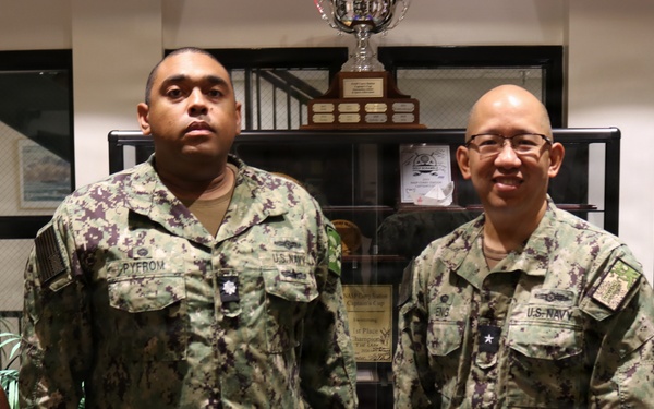Two U.S. Navy personnel in Navy Working Uniform (NWU Type III) camouflage are posing together indoors. The individual on the left has a name tape reading 'PYFROM' and the one on the right appears to have a name tape reading 'ENG', both displaying 'U.S. NAVY' chest tapes. They are standing in what appears to be a military office or lobby area with trophies and display cases visible in the background.
