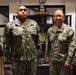 Two U.S. Navy personnel in Navy Working Uniform (NWU Type III) camouflage are posing together indoors. The individual on the left has a name tape reading 'PYFROM' and the one on the right appears to have a name tape reading 'ENG', both displaying 'U.S. NAVY' chest tapes. They are standing in what appears to be a military office or lobby area with trophies and display cases visible in the background.