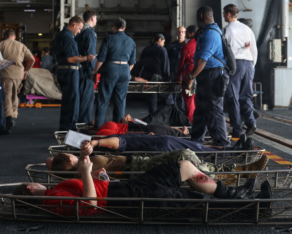 The image depicts what appears to be a military mass casualty (MASCAL) training exercise, likely aboard a naval vessel such as an aircraft carrier or amphibious ship. Multiple personnel in dark blue Navy coveralls are attending to simulated casualties lying on wire mesh stretchers/litters on the deck. Simulated wound moulage (fake blood/injuries) is visible on the 'casualties,' who are wearing red shirts to identify their role-player status.