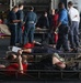 The image depicts what appears to be a military mass casualty (MASCAL) training exercise, likely aboard a naval vessel such as an aircraft carrier or amphibious ship. Multiple personnel in dark blue Navy coveralls are attending to simulated casualties lying on wire mesh stretchers/litters on the deck. Simulated wound moulage (fake blood/injuries) is visible on the 'casualties,' who are wearing red shirts to identify their role-player status.