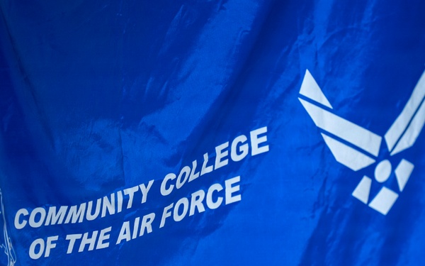 The image shows a blue flag belonging to the Community College of the Air Force (CCAF). The flag displays the institution's name in white text, along with the CCAF shield emblem on the left and the U.S. Air Force symbol (stylized wings) on the upper right. The flag appears to be hanging indoors, slightly draped and wrinkled.