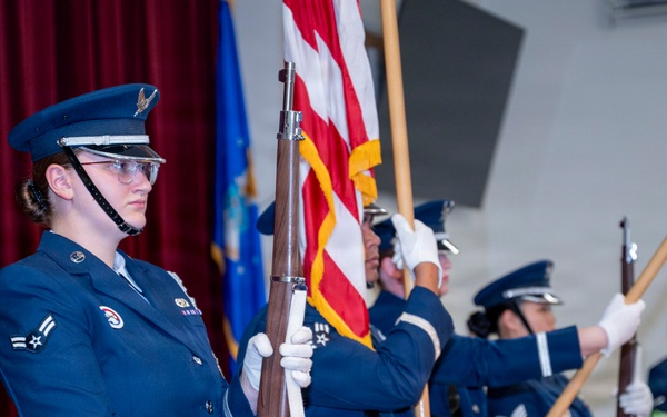 A United States Air Force color guard is performing a formal ceremony in an indoor venue, likely a base auditorium or gymnasium. Personnel in dress blue uniforms are carrying the American flag, a service flag, and ceremonial rifles with white gloves. A shield/emblem is visible on a podium in the background, suggesting an official military event such as a change of command or awards ceremony.