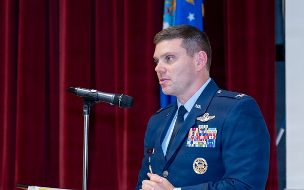 A U.S. Air Force officer in dress blue uniform is speaking at a podium during an official military ceremony or event. The officer displays multiple service ribbons, a pilot badge, and command insignia on his uniform. A unit shield/emblem is visible on the podium front, and a U.S. Air Force flag is visible in the background.