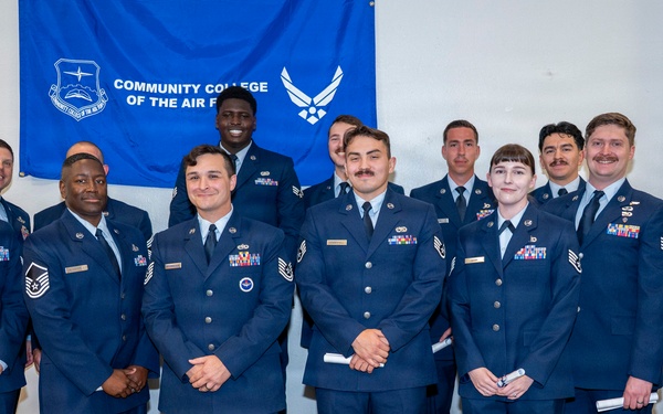 A group of approximately 13 United States Air Force personnel pose for a formal group photograph in front of a blue 'Community College of the Air Force' banner. The individuals are wearing Air Force Service Dress uniforms with various ranks and ribbons visible. This appears to be a graduation or academic achievement ceremony.