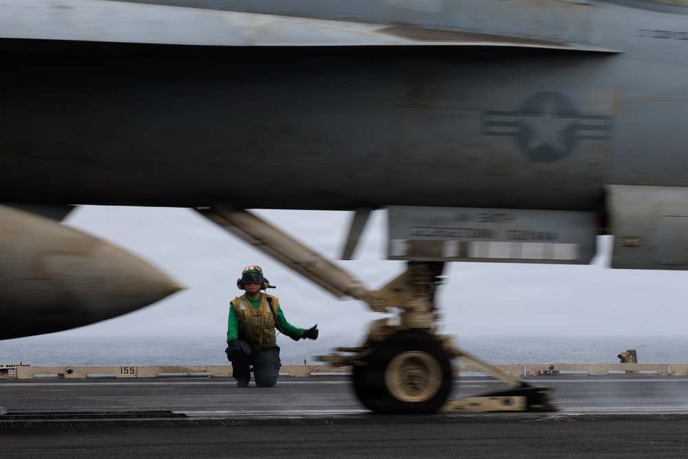 A U.S. Navy flight deck crew member in a green jersey and yellow safety vest kneels on an aircraft carrier flight deck, giving a thumbs-up signal beneath a military jet aircraft. The image is taken from a low angle, with the aircraft's fuselage and landing gear dominating the foreground in motion blur. The U.S. military roundel (star-and-bar insignia) is clearly visible on the aircraft's fuselage.