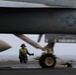 A U.S. Navy flight deck crew member in a green jersey and yellow safety vest kneels on an aircraft carrier flight deck, giving a thumbs-up signal beneath a military jet aircraft. The image is taken from a low angle, with the aircraft's fuselage and landing gear dominating the foreground in motion blur. The U.S. military roundel (star-and-bar insignia) is clearly visible on the aircraft's fuselage.