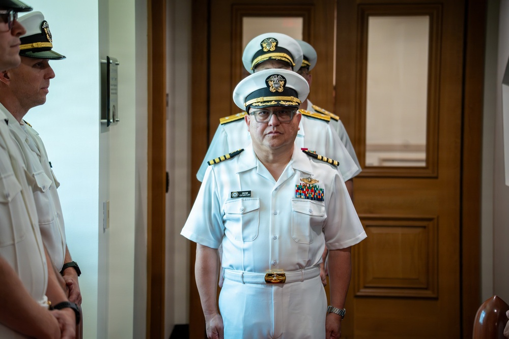 The image shows multiple U.S. Navy officers in white service dress uniforms standing in what appears to be an indoor military facility or office building. The central figure is a senior officer wearing a combination cover with gold braid, glasses, and multiple service ribbons on his chest. The scene appears to be a formal military ceremony or official event.