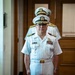 The image shows multiple U.S. Navy officers in white service dress uniforms standing in what appears to be an indoor military facility or office building. The central figure is a senior officer wearing a combination cover with gold braid, glasses, and multiple service ribbons on his chest. The scene appears to be a formal military ceremony or official event.