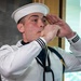A young male sailor in dress white uniform is playing a boatswain's pipe (bosun's whistle), a traditional naval instrument used for ceremonial calls and signaling. He is holding the small metal pipe to his lips with both hands raised in a characteristic playing position. The image appears to be taken indoors, likely during a formal naval ceremony or event.