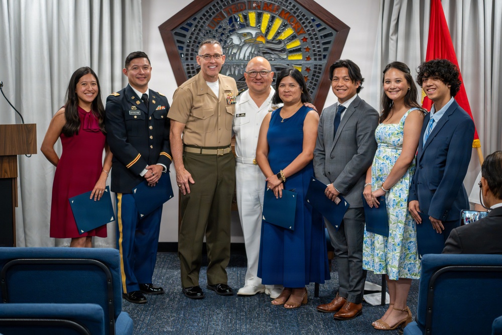 A group of eight people pose for a formal photograph in what appears to be a military ceremony room. The group includes personnel in U.S. Army dress uniform, U.S. Marine Corps service uniform, and U.S. Navy dress whites, alongside civilians in formal attire. Several individuals are holding blue folders, suggesting an awards or recognition ceremony.