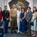 A group of eight people pose for a formal photograph in what appears to be a military ceremony room. The group includes personnel in U.S. Army dress uniform, U.S. Marine Corps service uniform, and U.S. Navy dress whites, alongside civilians in formal attire. Several individuals are holding blue folders, suggesting an awards or recognition ceremony.