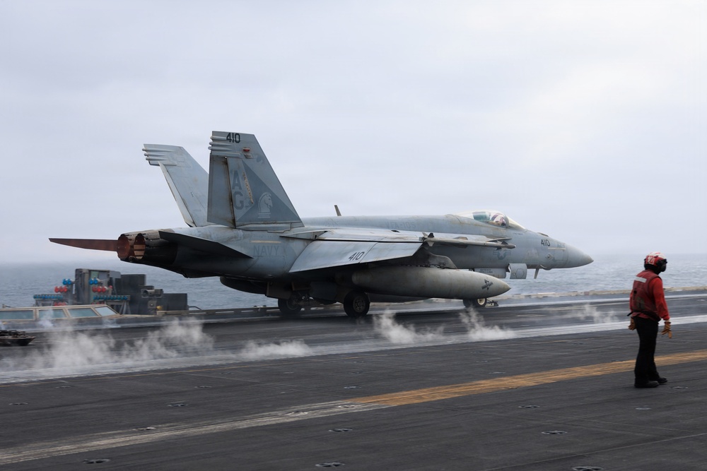 A U.S. Navy F/A-18 Hornet or Super Hornet bearing the modex number '410' is positioned on an aircraft carrier flight deck, appearing to be in the process of launching or preparing for catapult launch. Steam is visible rising from the catapult track beneath the aircraft. A flight deck crew member in a red jersey stands to the right observing the operation.