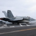 A U.S. Navy F/A-18 Hornet or Super Hornet bearing the modex number '410' is positioned on an aircraft carrier flight deck, appearing to be in the process of launching or preparing for catapult launch. Steam is visible rising from the catapult track beneath the aircraft. A flight deck crew member in a red jersey stands to the right observing the operation.