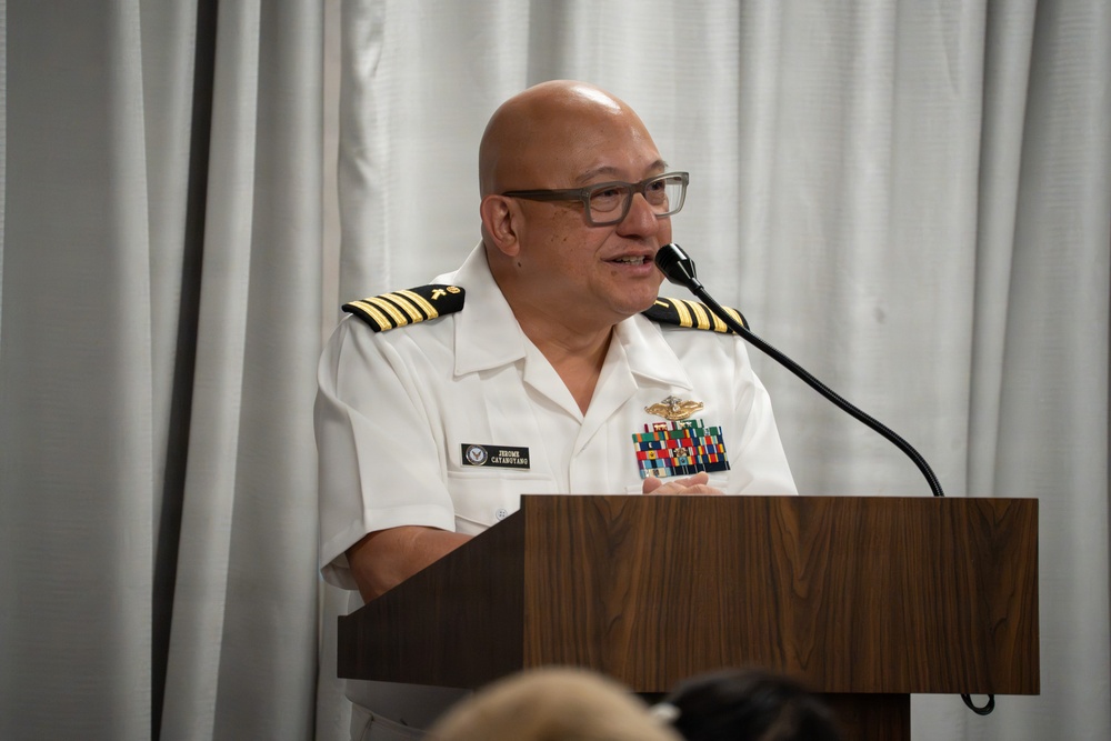 A U.S. Navy Captain in white service dress uniform is speaking at a wooden podium with a microphone. The officer's name tag reads 'CATABUTANO' with a first name appearing to begin with 'JEROME', and displays multiple service ribbons and qualification badges on the uniform. The setting appears to be an indoor ceremony or official function with white curtain backdrop.