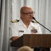 A U.S. Navy Captain in white service dress uniform is speaking at a wooden podium with a microphone. The officer's name tag reads 'CATABUTANO' with a first name appearing to begin with 'JEROME', and displays multiple service ribbons and qualification badges on the uniform. The setting appears to be an indoor ceremony or official function with white curtain backdrop.
