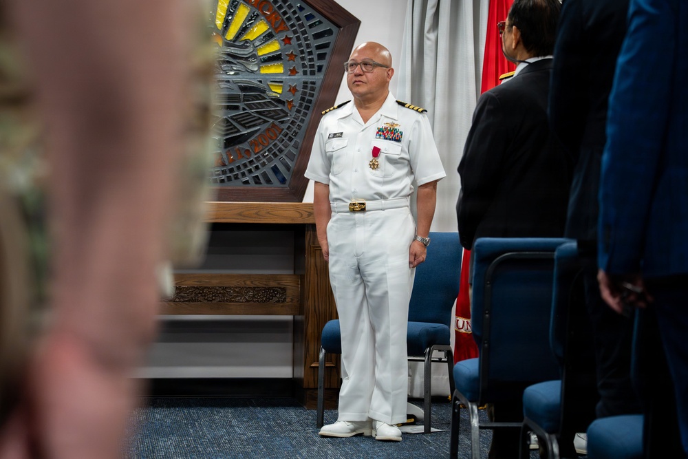 A U.S. Navy officer in dress white uniform stands at attention during what appears to be a formal military ceremony. He is wearing multiple service ribbons and a medal, suggesting this may be an awards or change of command ceremony. The setting appears to be an indoor military facility with a decorative wooden plaque and a red military flag visible in the background.
