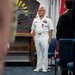 A U.S. Navy officer in dress white uniform stands at attention during what appears to be a formal military ceremony. He is wearing multiple service ribbons and a medal, suggesting this may be an awards or change of command ceremony. The setting appears to be an indoor military facility with a decorative wooden plaque and a red military flag visible in the background.
