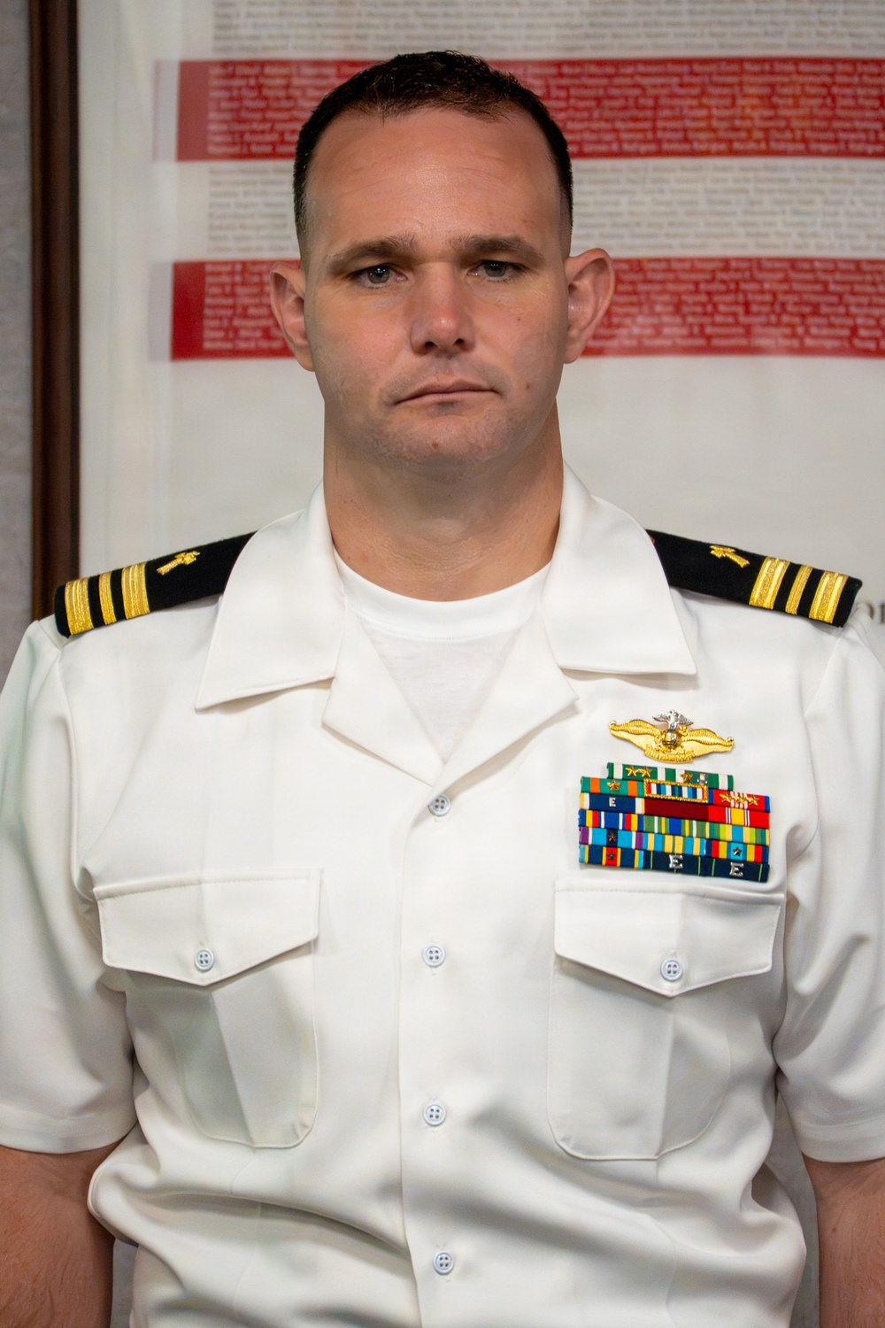 A U.S. Navy officer in Service Dress White uniform is photographed in a formal portrait setting. The officer displays multiple ribbons, medals, and a Naval Aviator or Naval Flight Officer breast insignia on the left chest. The background features what appears to be a memorial or honor roll display with names printed in red and black text.