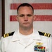 A U.S. Navy officer in Service Dress White uniform is photographed in a formal portrait setting. The officer displays multiple ribbons, medals, and a Naval Aviator or Naval Flight Officer breast insignia on the left chest. The background features what appears to be a memorial or honor roll display with names printed in red and black text.