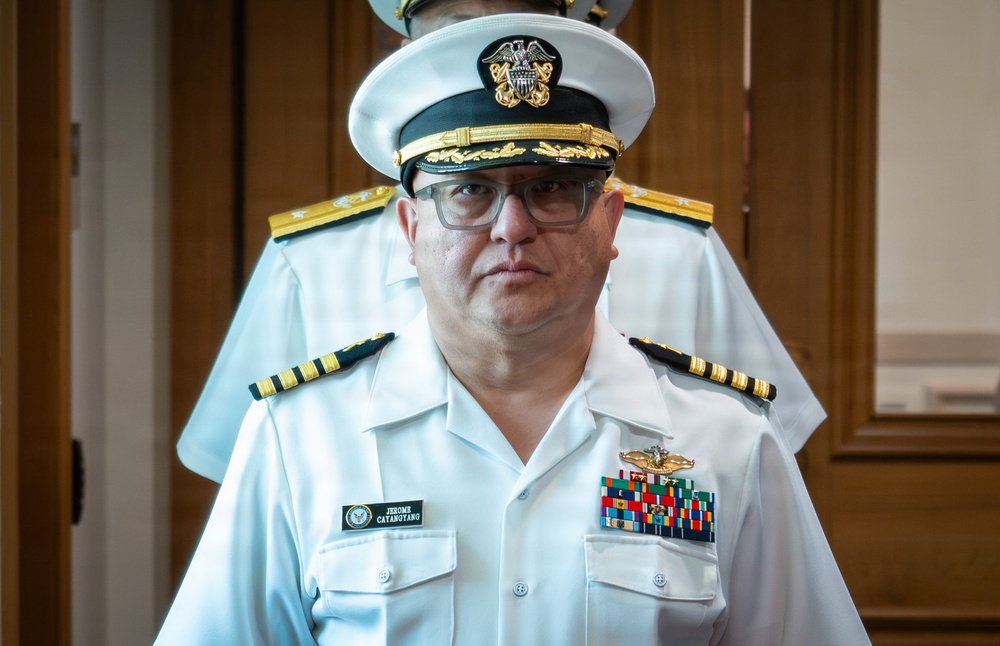 A U.S. Navy officer in white service dress uniform is photographed in what appears to be an indoor formal setting. His name tag reads 'Jerome Catabutano' and he displays multiple service ribbons and a naval aviator or warfare insignia on his chest. His rank insignia on the shoulder boards indicates a senior officer grade.