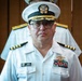 A U.S. Navy officer in white service dress uniform is photographed in what appears to be an indoor formal setting. His name tag reads 'Jerome Catabutano' and he displays multiple service ribbons and a naval aviator or warfare insignia on his chest. His rank insignia on the shoulder boards indicates a senior officer grade.