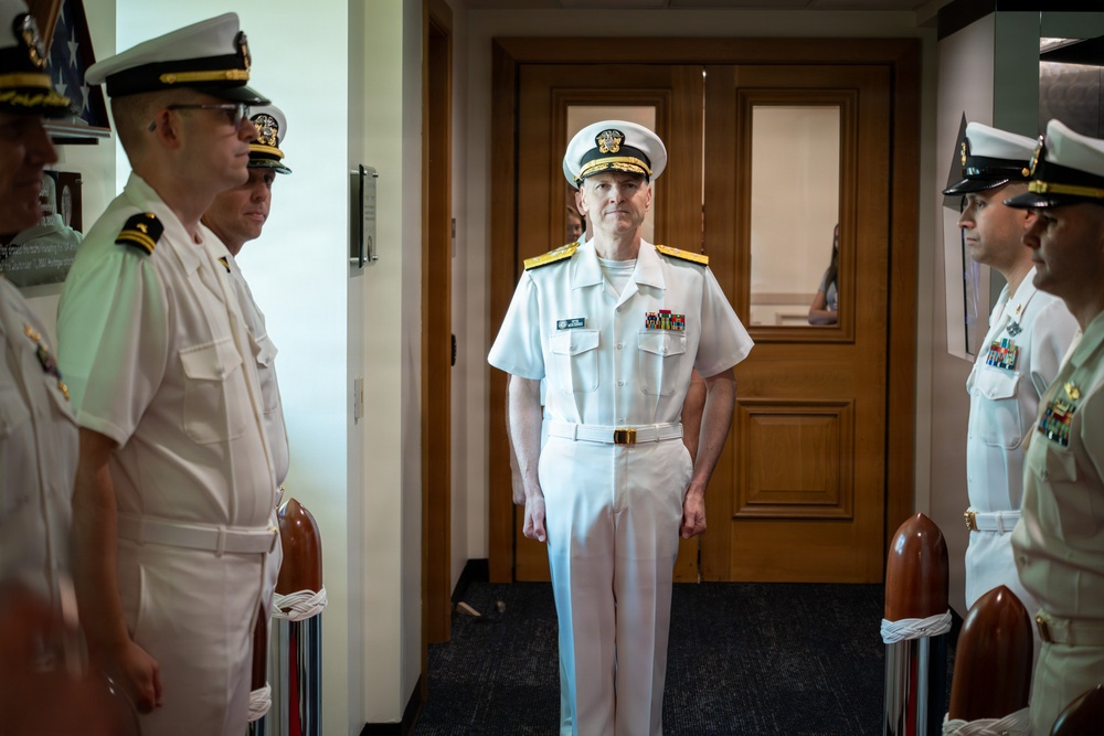 A senior U.S. Navy officer, identifiable by gold shoulder boards and multiple service ribbons, stands centered in a doorway while flanked by junior officers in white dress uniforms. The setting appears to be an indoor military facility, likely during a formal ceremony or official event. The composition suggests the central figure is being received or reviewed by the assembled personnel.