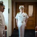 A senior U.S. Navy officer, identifiable by gold shoulder boards and multiple service ribbons, stands centered in a doorway while flanked by junior officers in white dress uniforms. The setting appears to be an indoor military facility, likely during a formal ceremony or official event. The composition suggests the central figure is being received or reviewed by the assembled personnel.