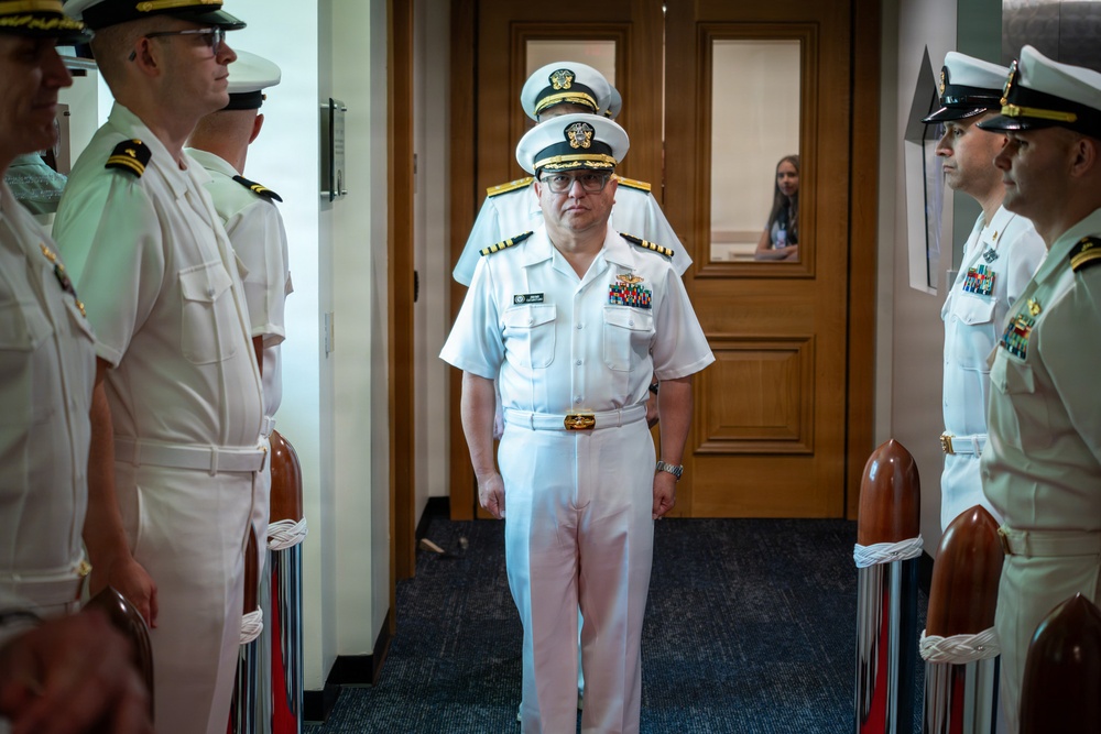 A senior U.S. Navy officer in white dress uniform walks through a ceremonial sideboy formation in an indoor corridor. The central figure displays multiple service ribbons and gold shoulder boards indicating flag or senior officer rank. The formal arrangement of personnel holding ceremonial swords or batons suggests an official military ceremony such as a change of command or retirement.