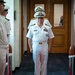 A senior U.S. Navy officer in white dress uniform walks through a ceremonial sideboy formation in an indoor corridor. The central figure displays multiple service ribbons and gold shoulder boards indicating flag or senior officer rank. The formal arrangement of personnel holding ceremonial swords or batons suggests an official military ceremony such as a change of command or retirement.