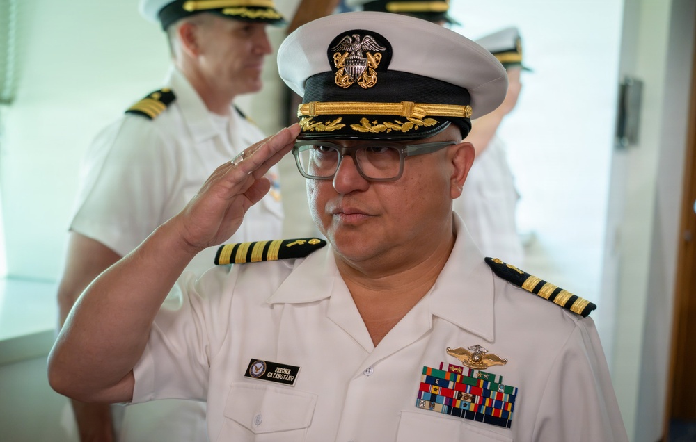 A U.S. Navy officer is rendering a salute while wearing a white service dress uniform with a combination cover. His name tag reads 'Jerome Catamotang' and he displays multiple service ribbons, a surface warfare insignia, and captain-level rank insignia. A second Navy officer in the background also appears to be saluting.